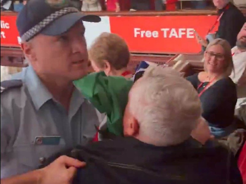 A uniformed police officer in a peaked cap stands among a crowd of civilians, with a red "Free TAFE" banner visible in the background.