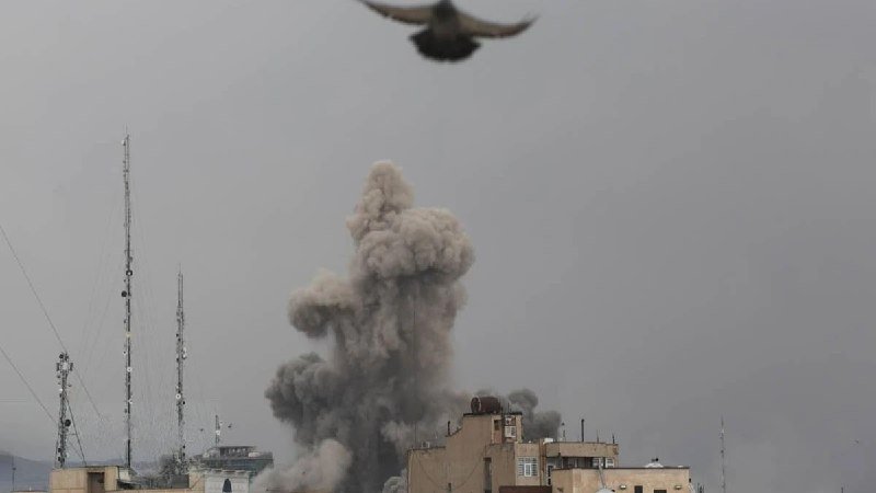 A large plume of gray smoke billows upward from behind rooftops and communication towers against a hazy sky, with a bird flying overhead.