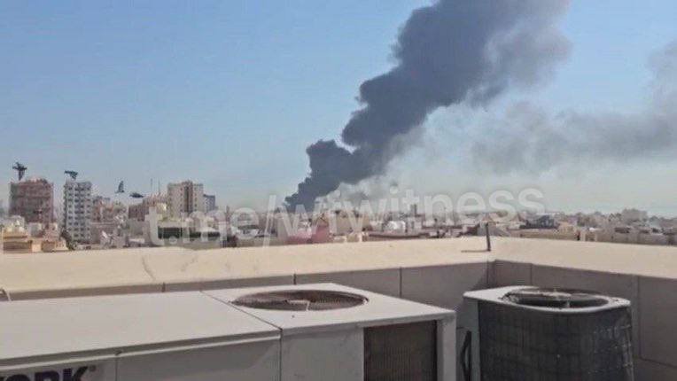 Thick black smoke rises over a desert cityscape behind rooftop AC units and a low parapet wall, with a "theonlywitness" watermark across the center.
