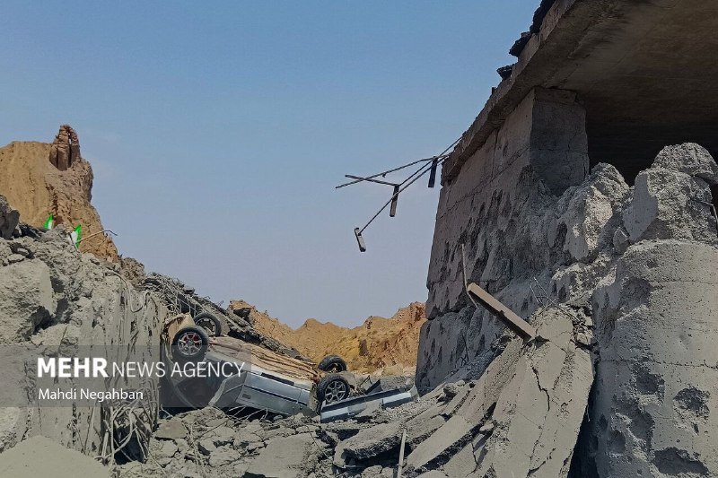 An overturned car lies among rubble and debris beside a damaged concrete structure under a clear sky.