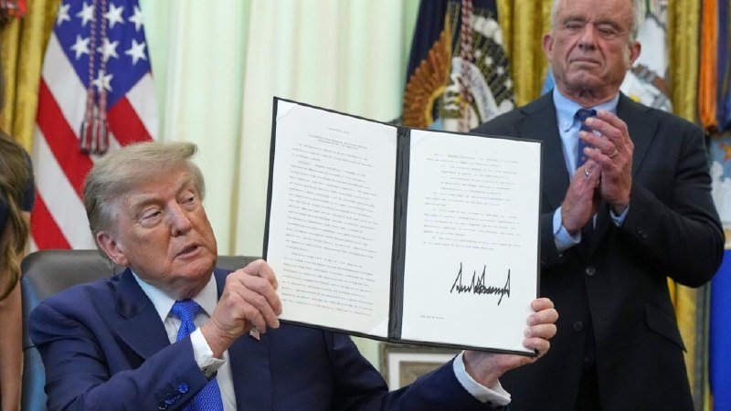 A man in a blue suit displays an open, signed document while another man in a dark suit applauds, with American flags visible behind them.