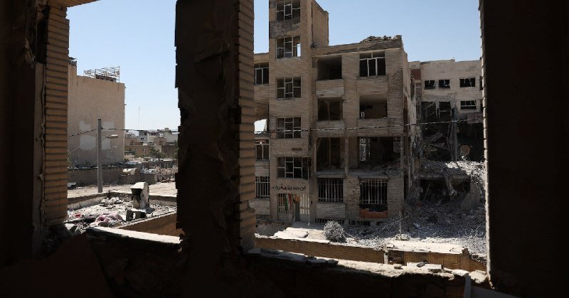 Heavily damaged multi-story buildings with shattered windows and rubble-strewn debris surround a courtyard, viewed through a broken wall opening under clear skies.