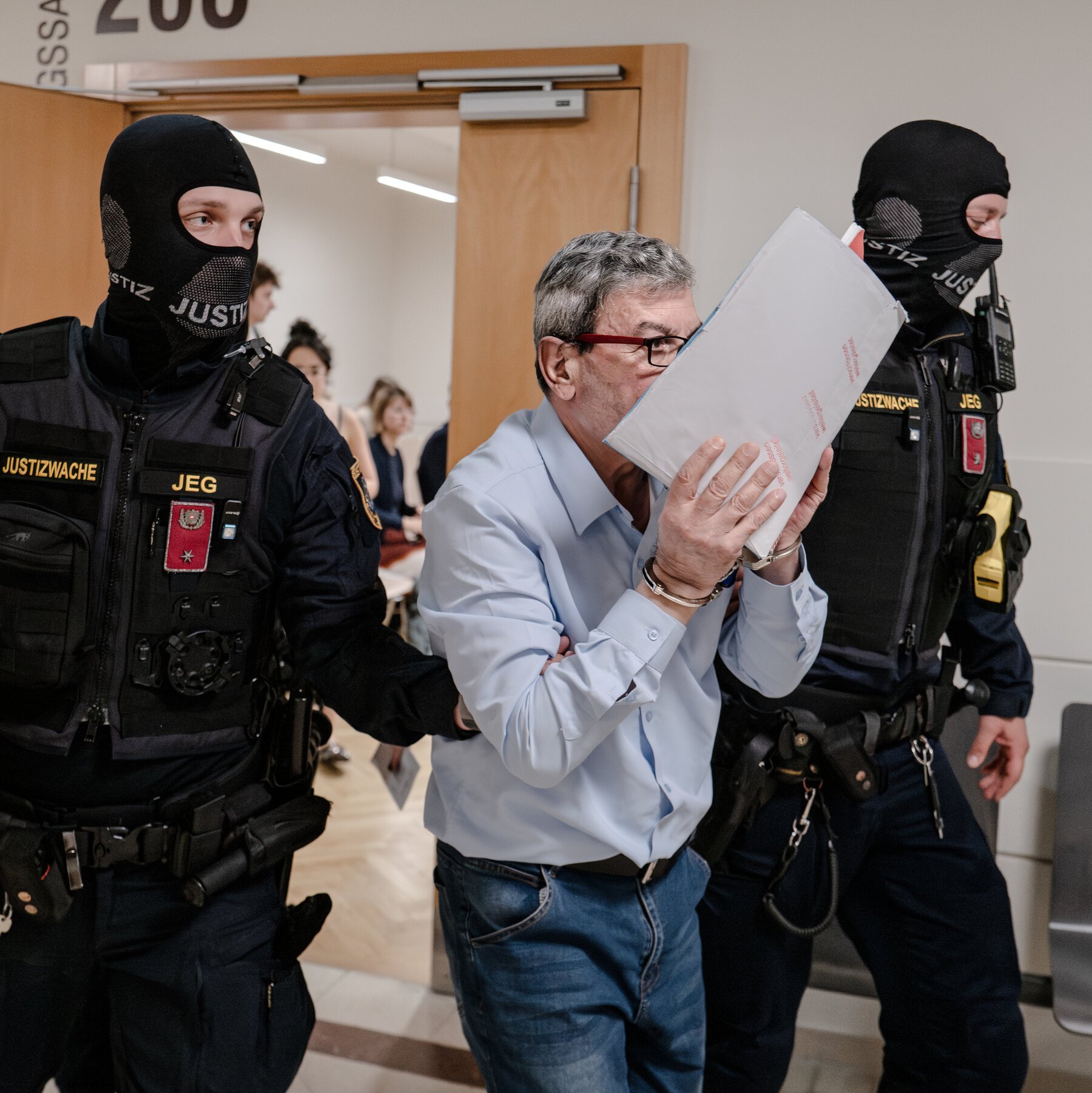 A handcuffed man in a blue shirt holds an envelope over his face while being escorted by two masked officers marked "JUSTIZWACHE JEG" through a doorway.