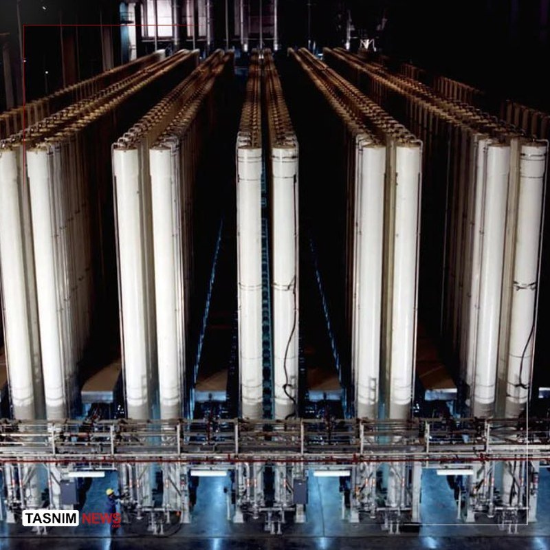 Rows of tall, white cylindrical industrial equipment stand in a dimly lit facility, with a worker in a hard hat visible at the bottom left.