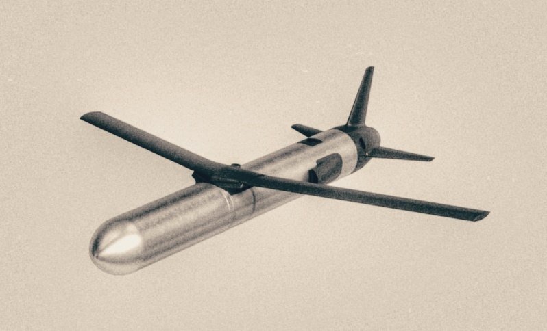 A military missile with a pointed nose, long swept wings, and rear tail fins is suspended against a plain light background.