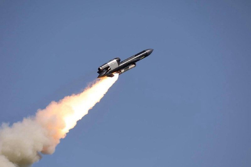 A rocket with a smaller craft mounted on top ascends diagonally against a clear blue sky, propelled by a bright orange flame and trailing thick white exhaust.