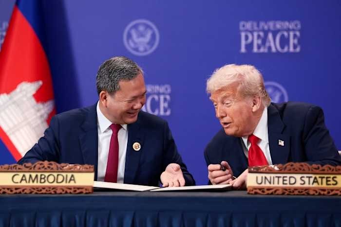 Two men in dark suits sit at a table with nameplates reading "CAMBODIA" and "UNITED STATES," conversing in front of a blue backdrop displaying "DELIVERING PEACE."