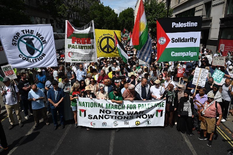 A large crowd marches down a city street holding banners reading "Stop the War Coalition," "Free Palestine," "Palestine Solidarity Campaign," and "National March for Palestine: No to War, Sanctions Now."