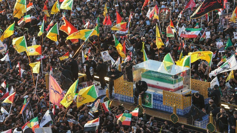 A large crowd waves yellow, red, and green flags while surrounding a glass-encased coffin draped in the Iranian flag during a public procession.
