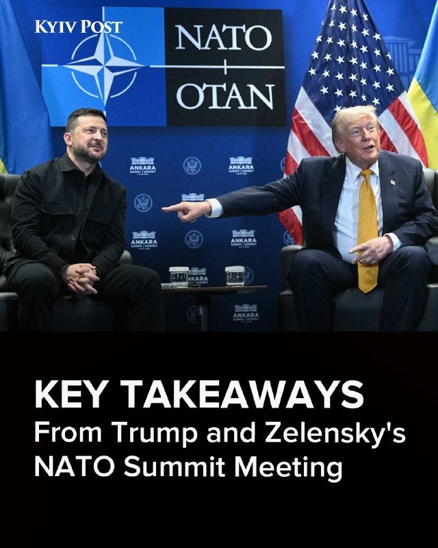 Two men sit seated in chairs before a NATO backdrop and national flags, with one man in a dark suit pointing toward the other.