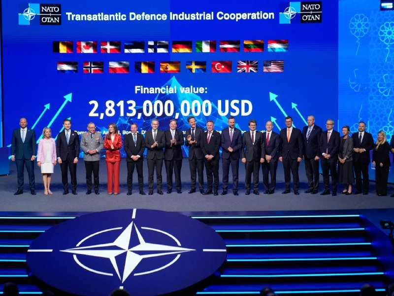 Officials stand together on stage with NATO logo, beneath a screen displaying "Transatlantic Defence Industrial Cooperation" and a financial value of 2,813,000,000 USD alongside member flags.