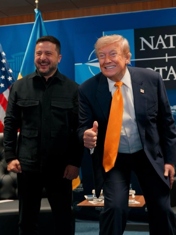 Two men—one in a dark jacket, the other in a navy suit with an orange tie giving a thumbs-up—smile in front of a NATO banner with Ukrainian and American flags nearby.