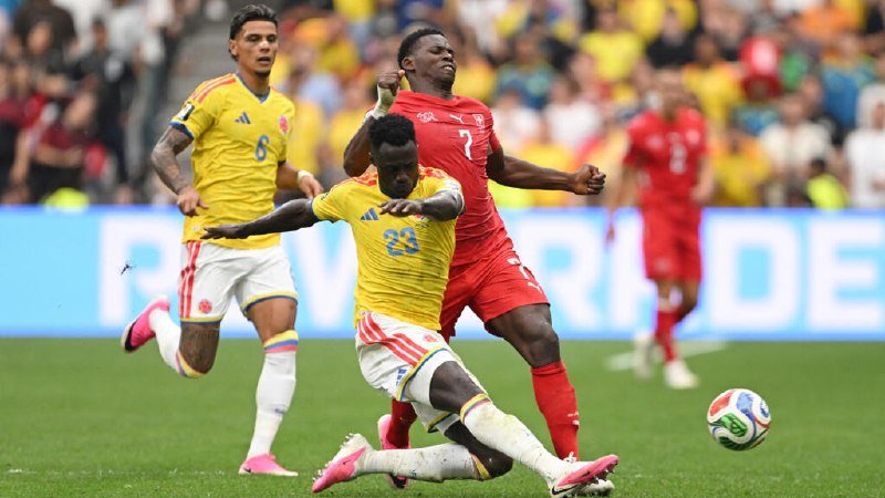 Two soccer players in yellow and red kits contest for the ball during a match, with a crowded stadium in the background.