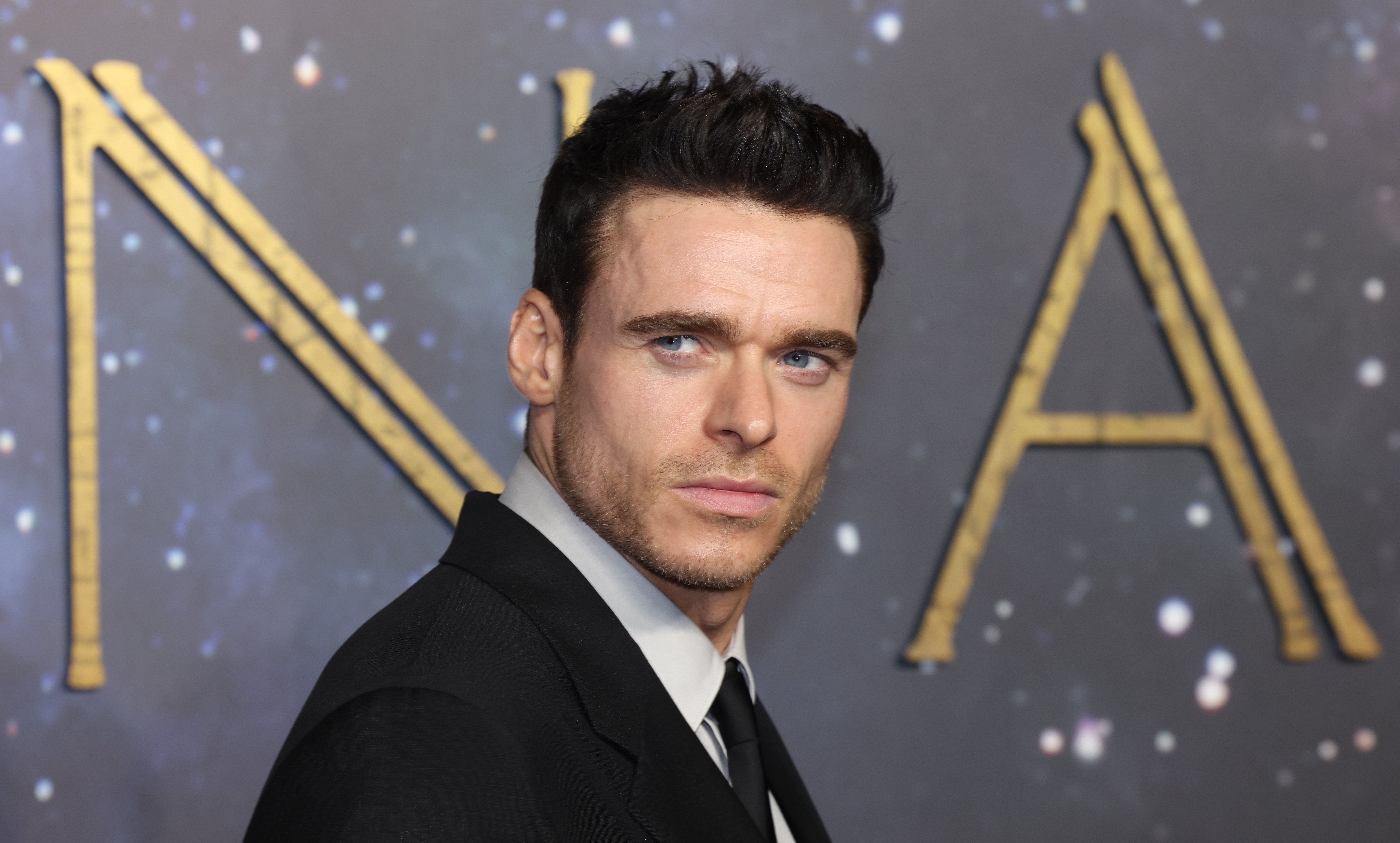 A dark-haired man in a black suit and tie poses at a starry backdrop displaying large gold letters "NA."