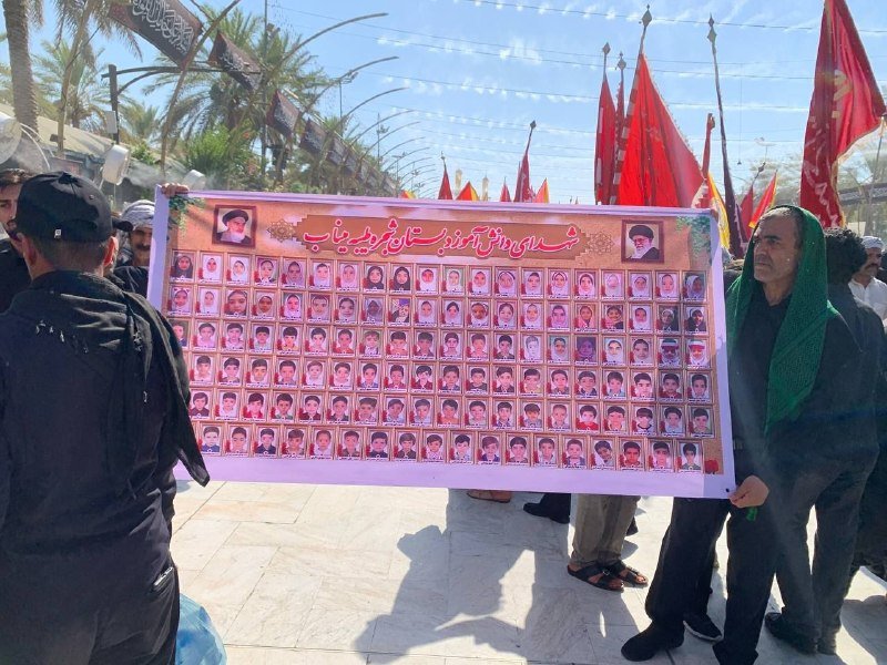 Demonstrators hold a large banner displaying rows of portrait photos and Arabic script, with red flags visible in the background on a paved street.
