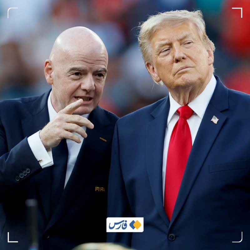 Two men in suits stand side by side, one bald in a dark suit gesturing with his hand, the other with blond hair wearing a red tie and American flag pin.