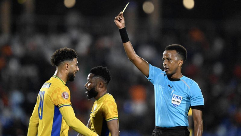 A referee in a light blue FIFA jersey holds up a yellow card while facing two soccer players wearing yellow and blue uniforms during a match.