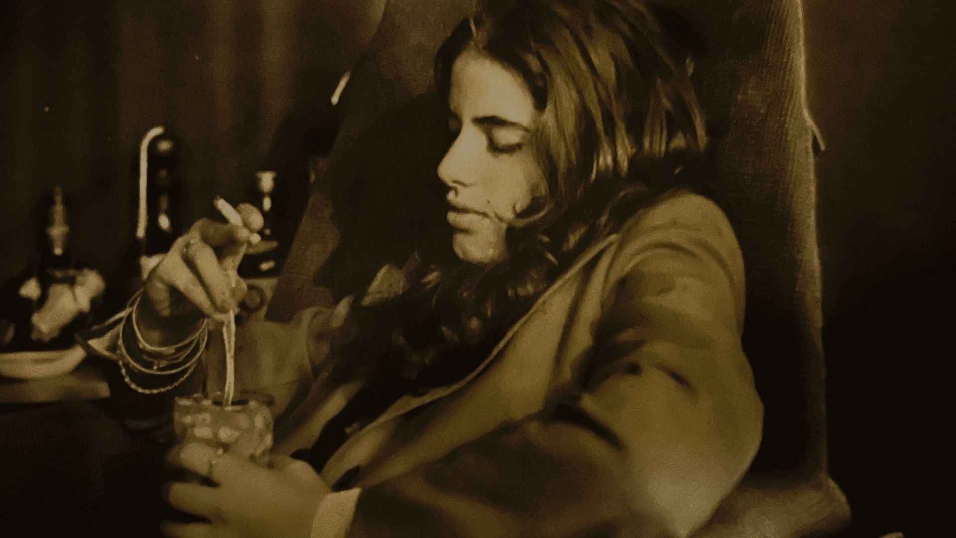 A woman with shoulder-length wavy hair sits in a wooden chair, manipulating an intricate figurine adorned with beaded strands in a dimly lit setting.