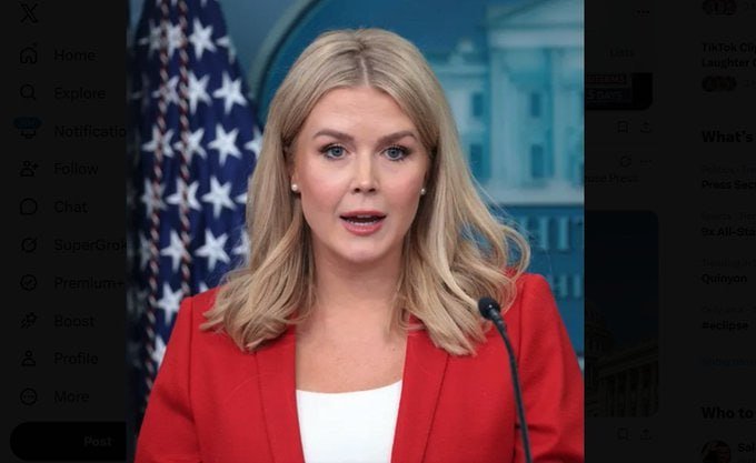 A blonde woman in a red blazer speaks at a podium with a microphone, an American flag and white building visible in the background.