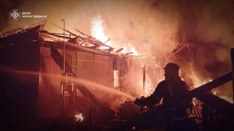A firefighter in silhouette sprays a water hose onto a large burning structure engulfed in flames and thick smoke at night, with a "ДСНС ЧЕРНІГІВЩИНИ" emblem in the corner.