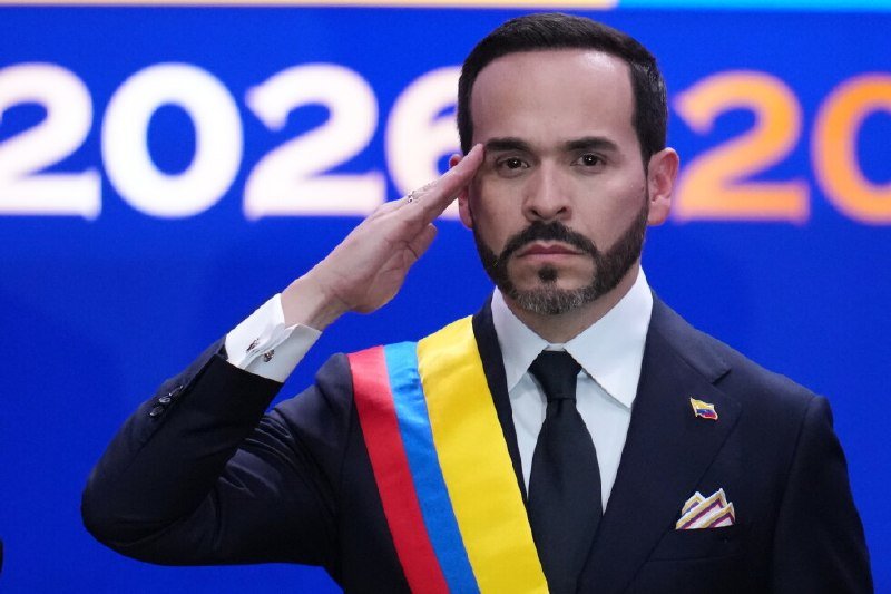 A bearded man in a dark suit wearing a yellow, blue, and red sash salutes before a blue backdrop displaying partial white and orange "2026" text.