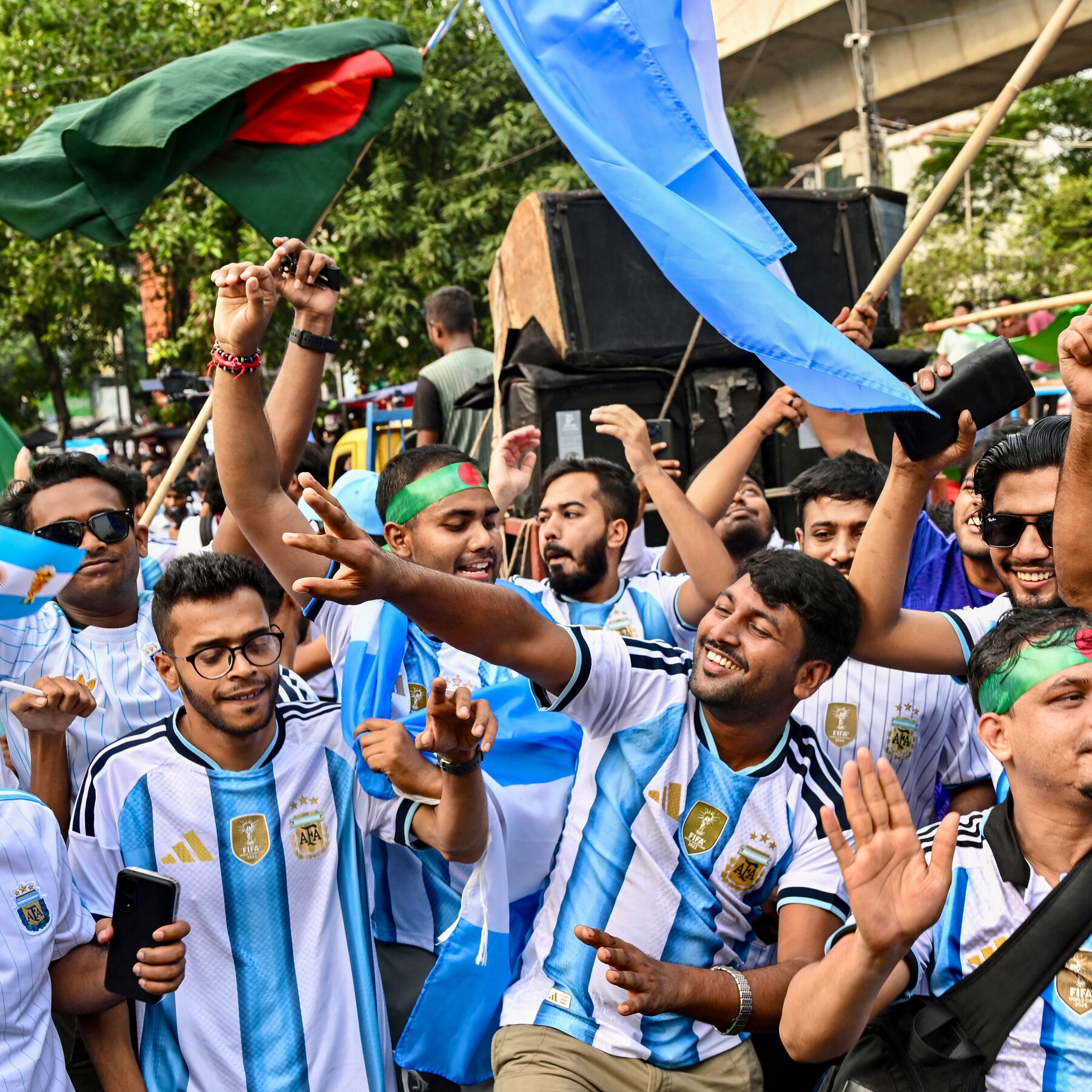 Crowds of fans in Argentina jerseys celebrate in a street, waving the green and red flag of Bangladesh and light blue flags.