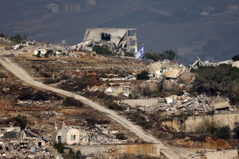 Israeli flag flies amid heavily destroyed buildings and rubble on a hillside with a dirt road winding through the debris.