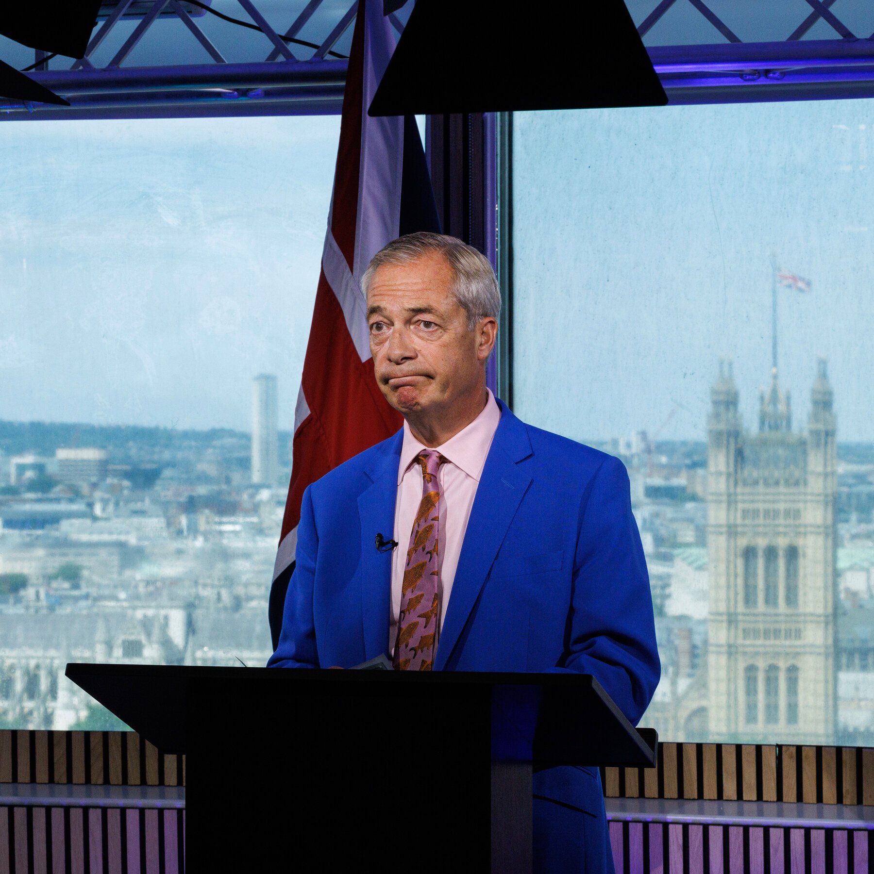 A grey-haired man in a blue suit and pink tie stands at a black podium, with a Union Jack flag and the Palace of Westminster visible through the window behind him.