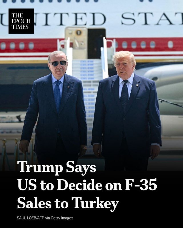 Two men in dark suits stand side by side in front of an aircraft with "UNITED STATES" lettering and a jet bridge visible behind them.