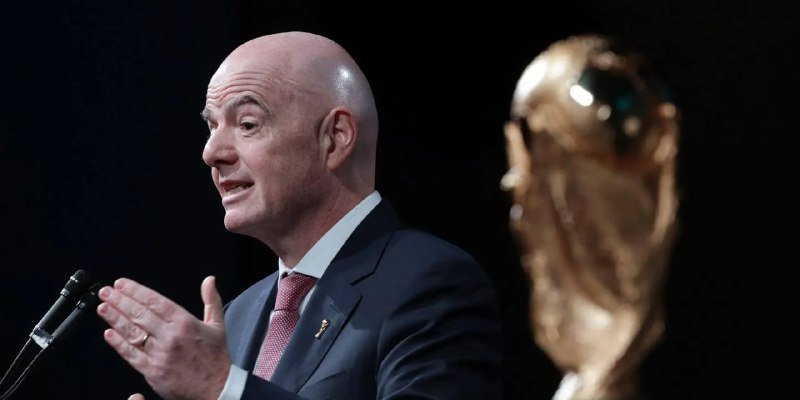 A bald man in a dark suit and red tie speaks at a microphone with his hands raised, standing beside a golden trophy against a dark background.