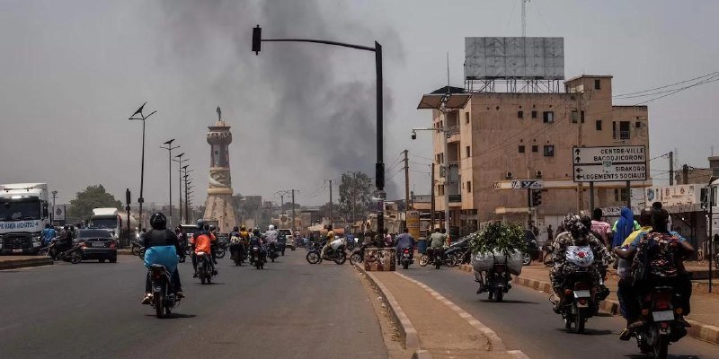 Motorcyclists and a truck travel along a busy urban roadway as thick black smoke rises near a tall monument and buildings with directional signs.