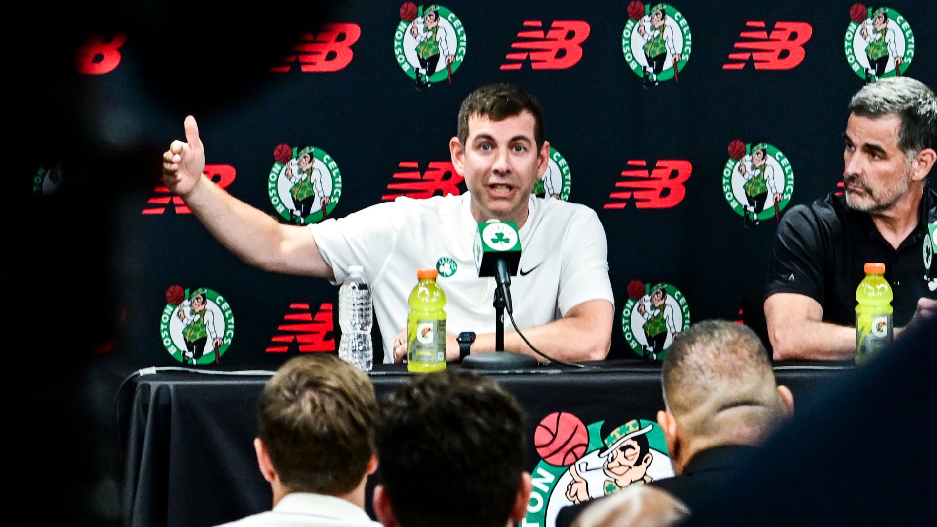 A man in a white Celtics shirt gestures while speaking at a press conference table, beside a gray-haired man in a dark shirt, against a Boston Celtics and New Balance backdrop.
