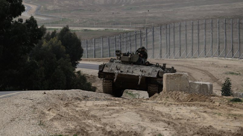 A military tank with a soldier visible in the turret drives along a dirt path beside a tall border fence in an arid landscape.