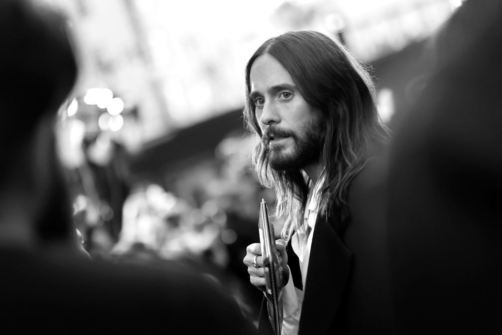 A long-haired, bearded man in a suit holds an award statuette while surrounded by a blurred crowd in this black-and-white photograph.