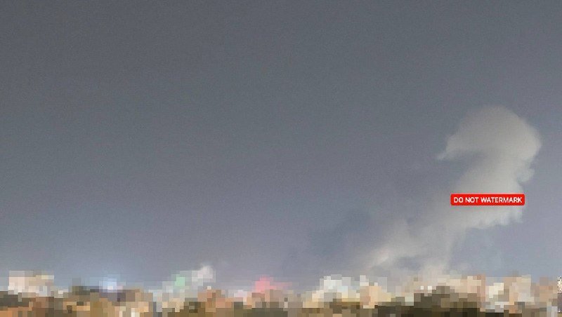 A night sky with a large white smoke plume rising on the right, above a heavily pixelated cityscape of glowing lights.