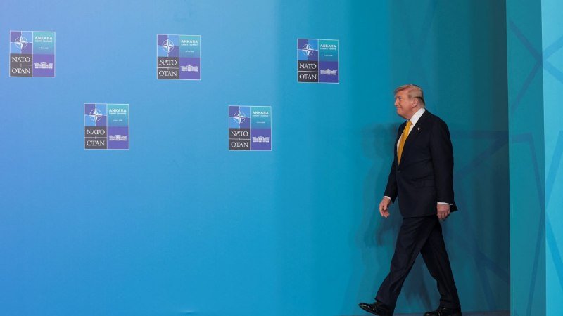 A man in a dark suit with a yellow tie walks past a turquoise wall displaying multiple "NATO Ankara Summit" logos.