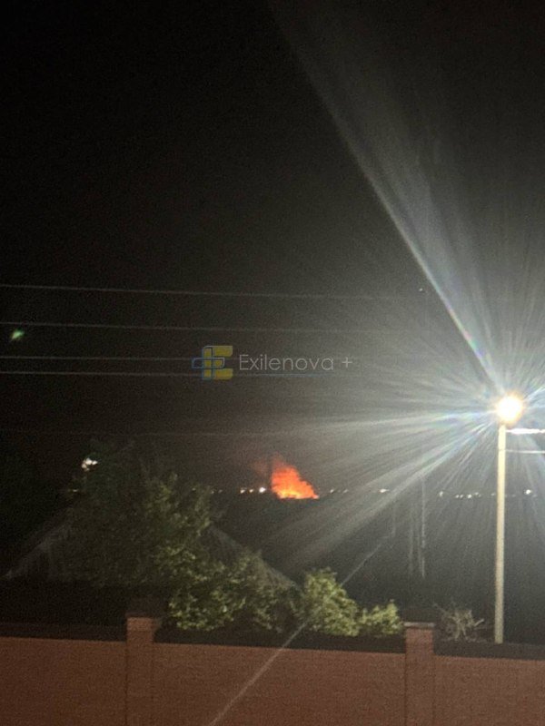 A nighttime photograph shows an orange glow from a fire on the horizon, contrasted against a bright streetlight and dark silhouettes of trees and rooftops in the foreground.