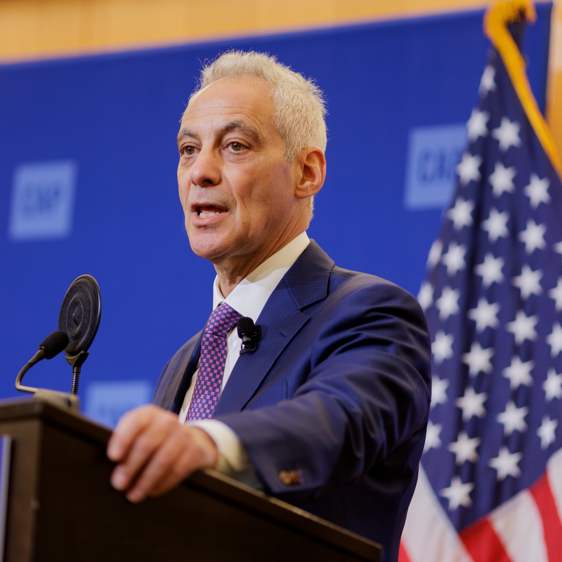 A man with white hair in a navy suit and patterned tie speaks at a podium with microphones, standing before a blue backdrop and an American flag.