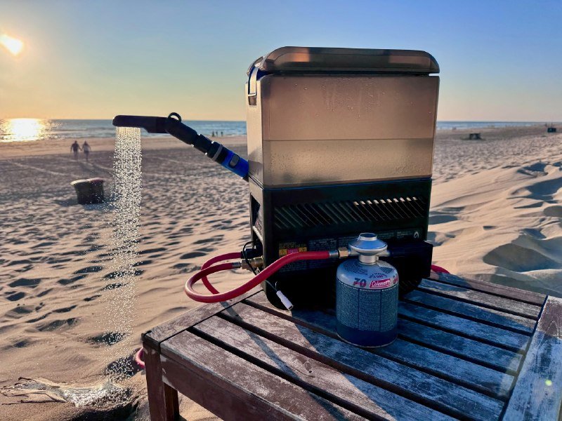 A portable gas-powered shower unit with an attached fuel canister sits on a wooden table on a sandy beach, spraying water from its nozzle.