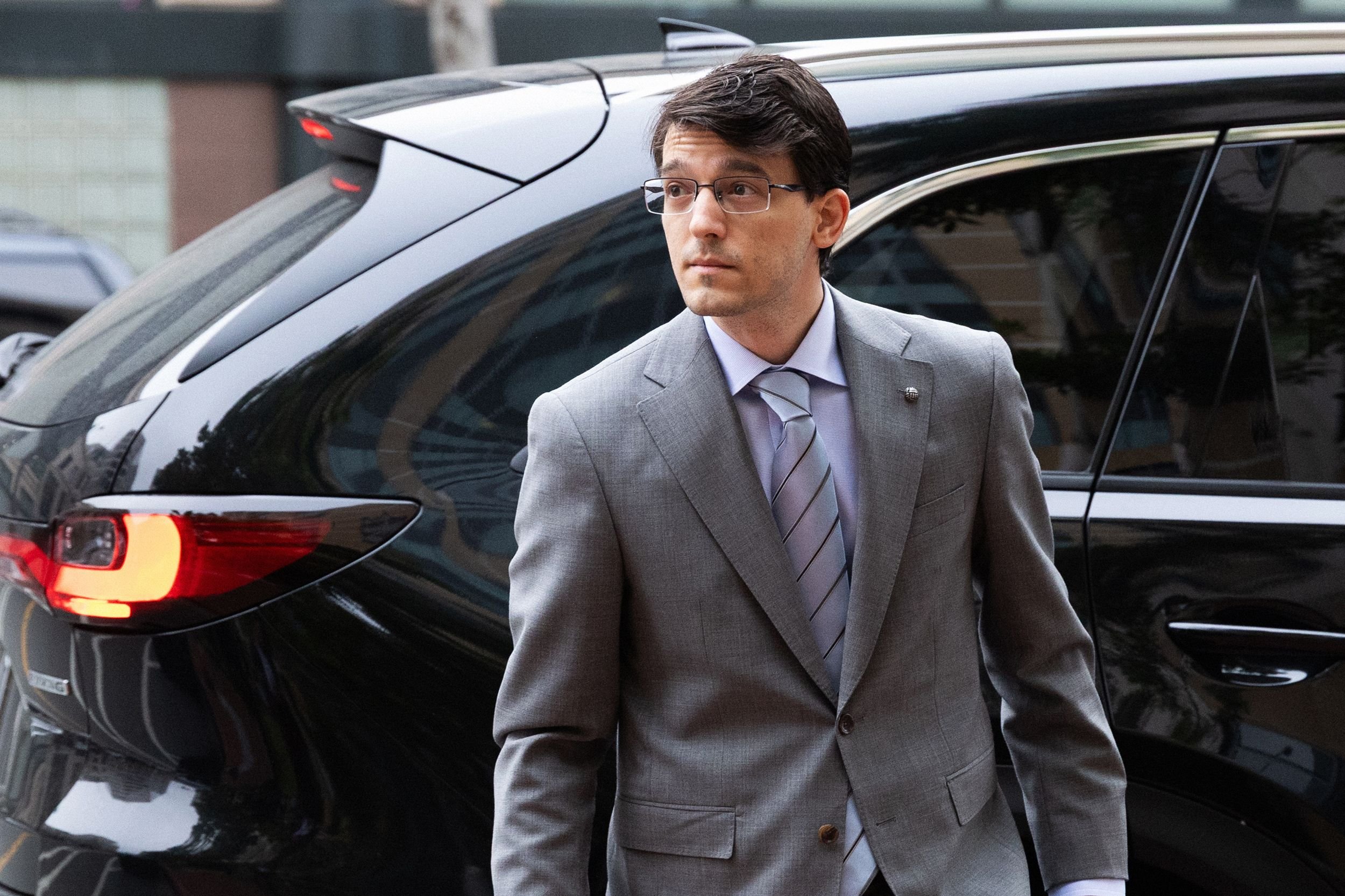 A man in a gray suit, lavender shirt, striped tie, and glasses walks past a black SUV with brake lights illuminated.