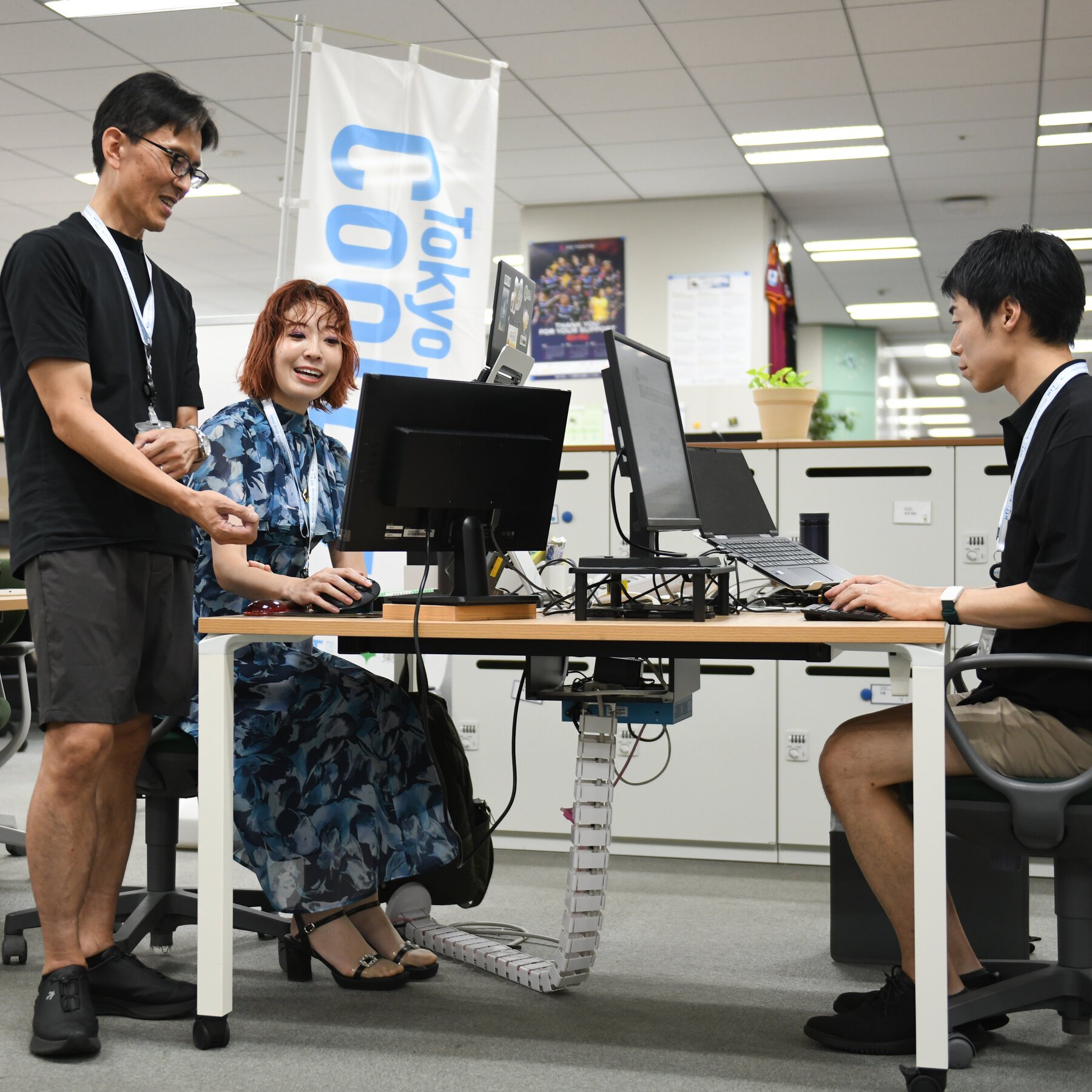 Three people gather around a desk with computer monitors in a brightly lit office space, with two seated working on devices while a man in a black shirt and shorts stands nearby smiling.