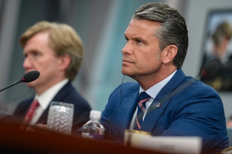 Two men in suits sit at a panel table with microphones, glasses, and a water bottle; one wears an earpiece while looking forward.