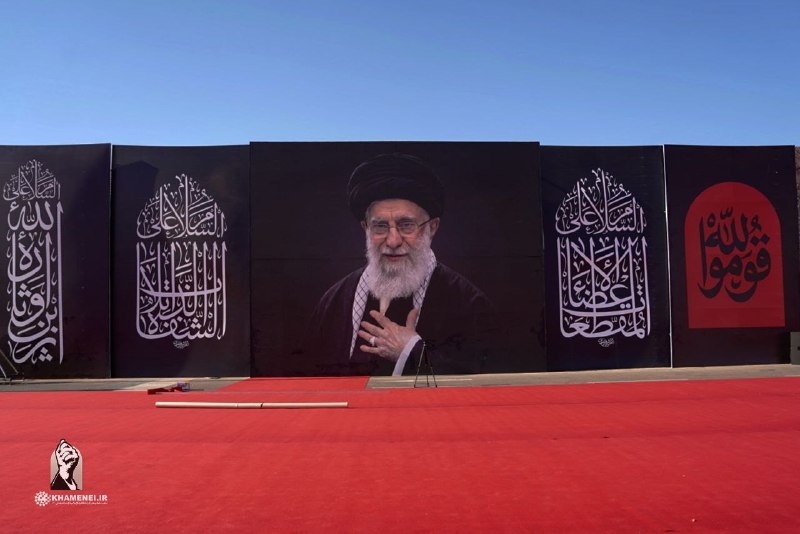 A bearded man wearing glasses, a black turban, and brown clerical robes stands beside a waving Iranian flag against a dark blue background.