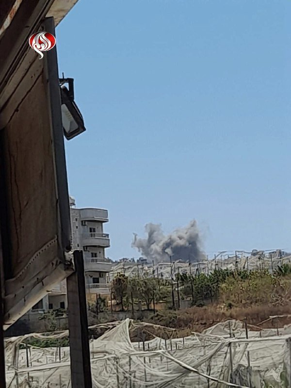 A plume of grey smoke rises over damaged greenhouses and a partially visible apartment building under a clear blue sky.