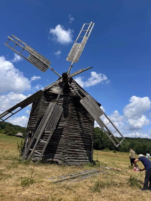 A weathered wooden windmill with four large sail frames stands in a grassy field under a partly cloudy sky, with two people near its base.