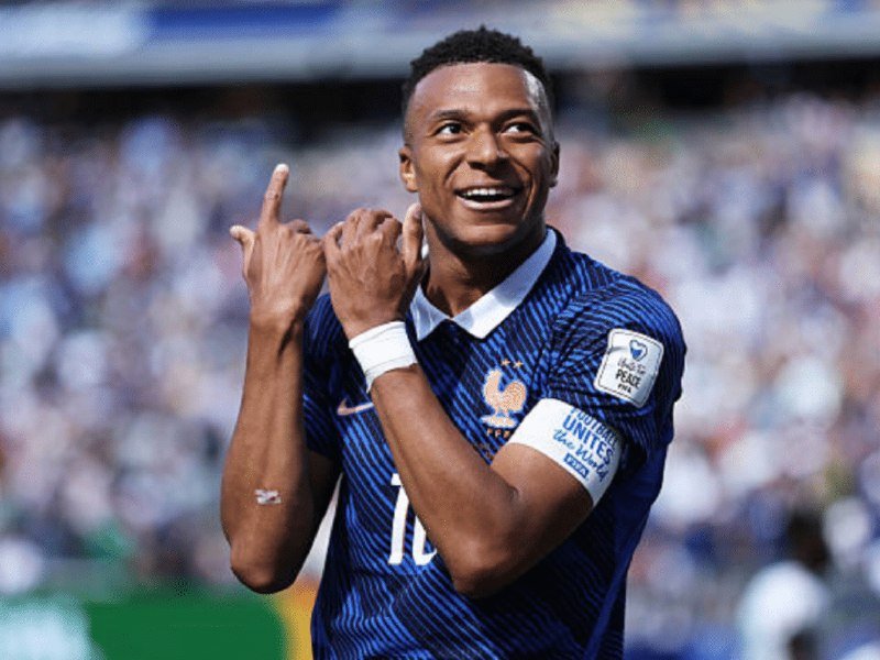 A soccer player in a blue France jersey smiles and points upward in celebration during a match before a stadium crowd.