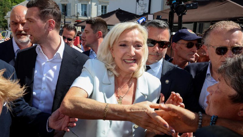 A smiling blonde woman in a white blazer shakes hands with supporters while being escorted by men in suits in a crowded outdoor setting.