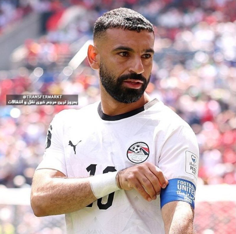 A bearded man in a white Puma jersey with a football badge adjusts a blue "Football Unites" captain's armband on his left bicep in a crowded stadium.