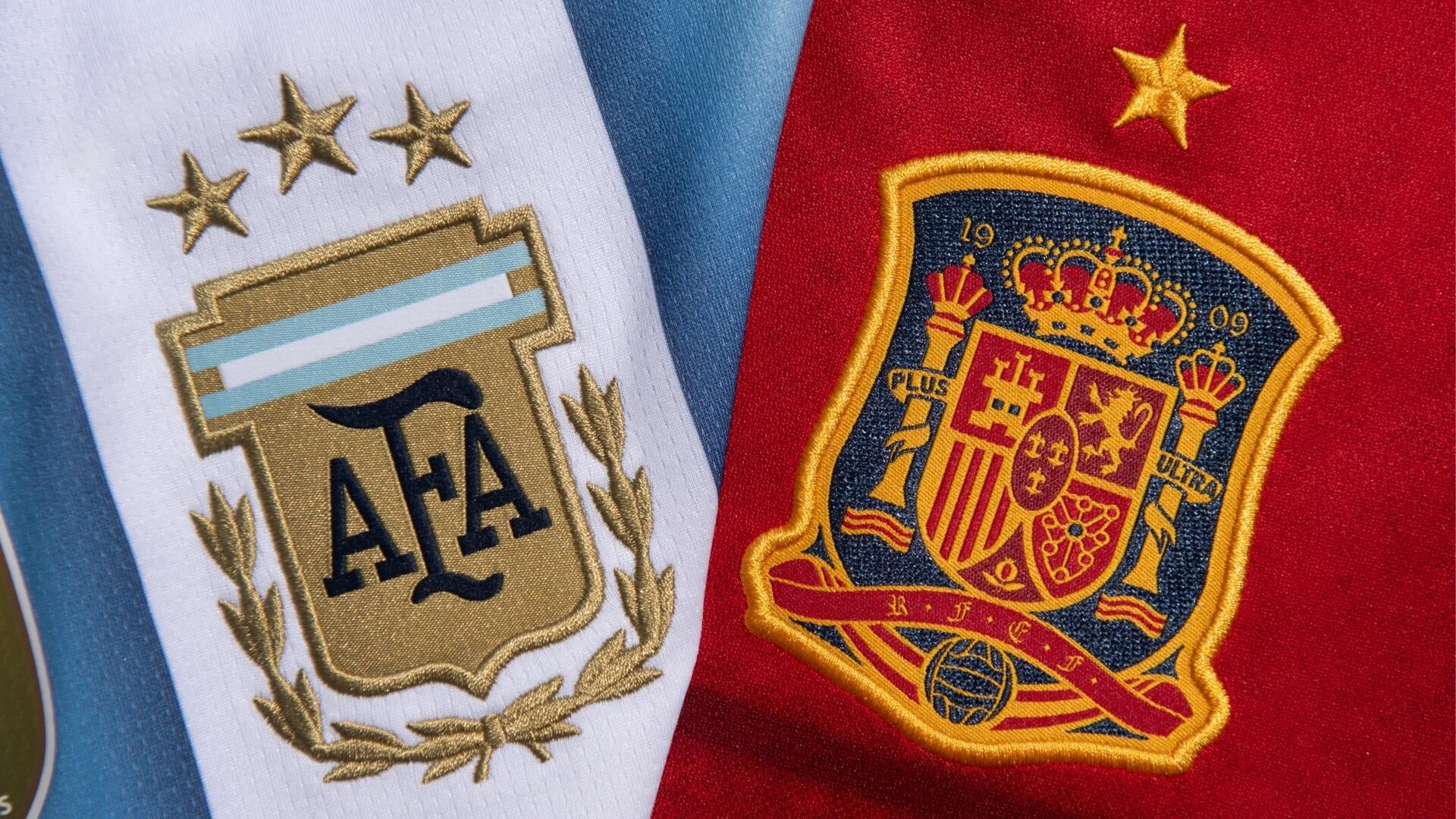 Close-up of two embroidered football jerseys side by side: a white and light-blue one featuring a gold "AFA" crest topped with stars, and a red one displaying a Spanish national team coat of arms with crown and "PLUS ULTRA" banner.