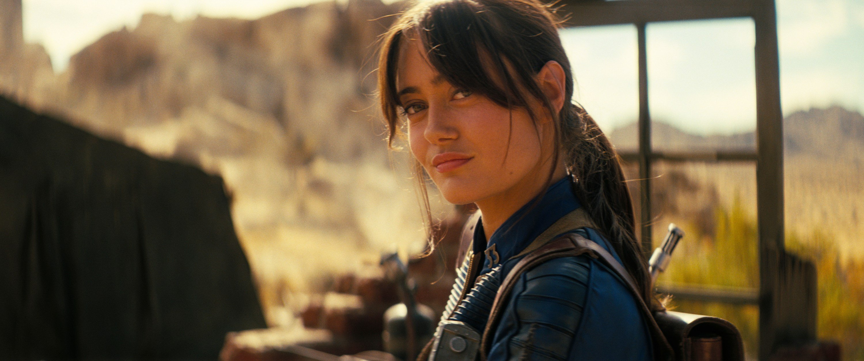 A young woman with dark hair in a ponytail, wearing a blue uniform with a corrugated tube, looks over her shoulder against a sunlit desert landscape.